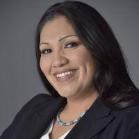 Text for Photo: A photo of a woman captured from the waist up, smiling warmly as she looks directly at the camera. She is an American Indian with long, brown hair, dressed in a white blouse and a black blazer, complemented by a striking turquoise necklace. Her arms rest naturally at her sides, projecting a confident and approachable demeanor.