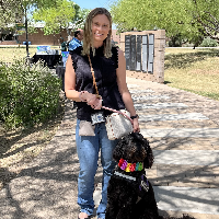  A white woman with shoulder length blonde hair wearing a black tank top, jeans, and black ballet flat shoes. A beige purse with a brown strap hangs from her right shoulder across to her left hip. She is holding a pink leash attached to a black fluffy dog sitting at her left leg with a service dog vest and rainbow collar. They are on a concrete and stone walkway with green bushes and trees, a wall with plaques, black tables, buildings, and grass behind them.