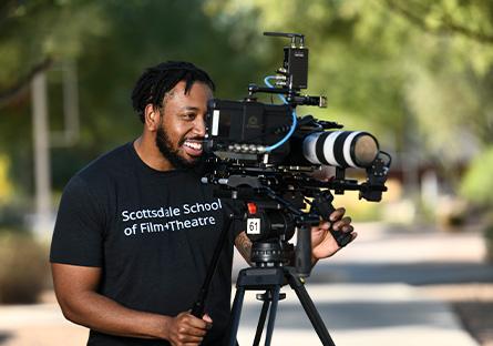 A smiling film school student is looking through camera lens. The student is wearing a Scottsdale Community College t-shirt.