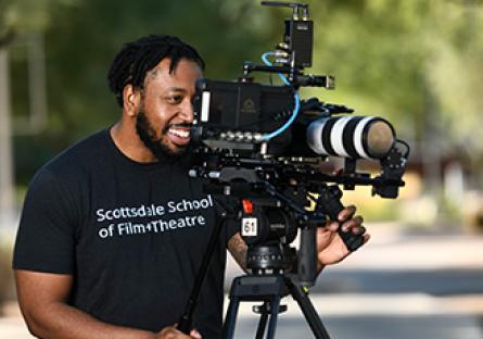 A smiling film school student is looking through camera lens. The student is wearing a Scottsdale Community College t-shirt.